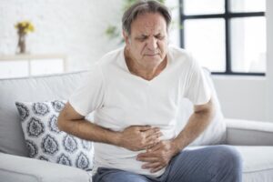 A senior man sitting on the sofa is suffering from severe constipation. He is holding his stomach with both hands
